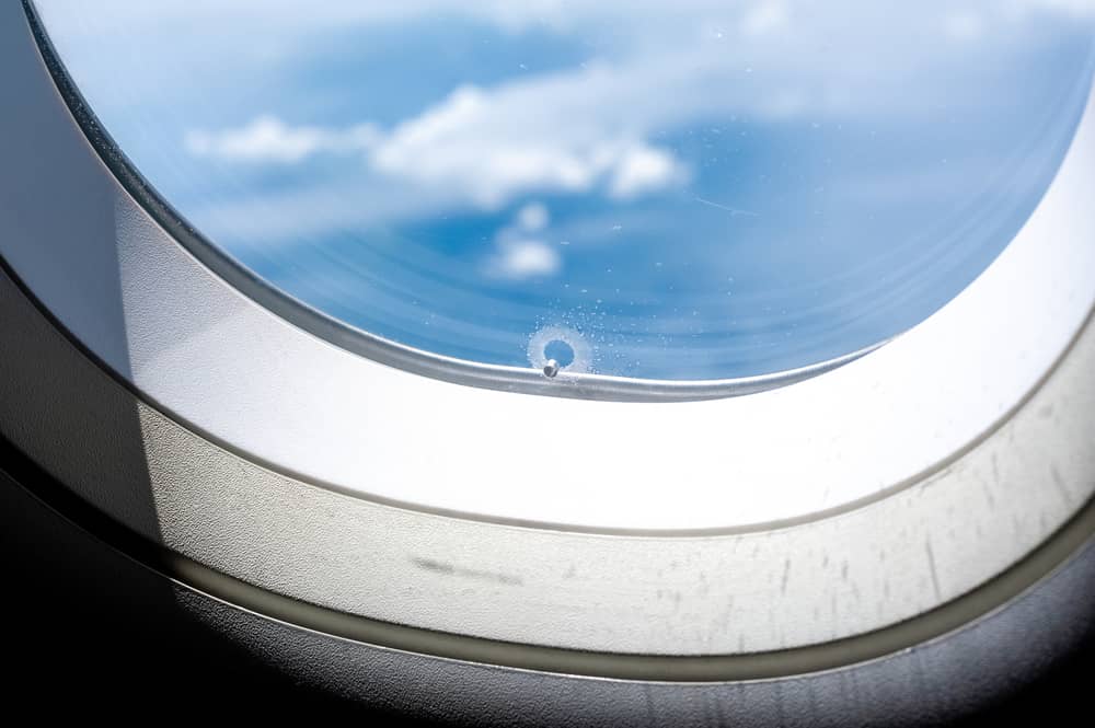 Primer plano de un pequeño agujero en la ventana de un avión, con un cielo azul y nubes visibles en el fondo.