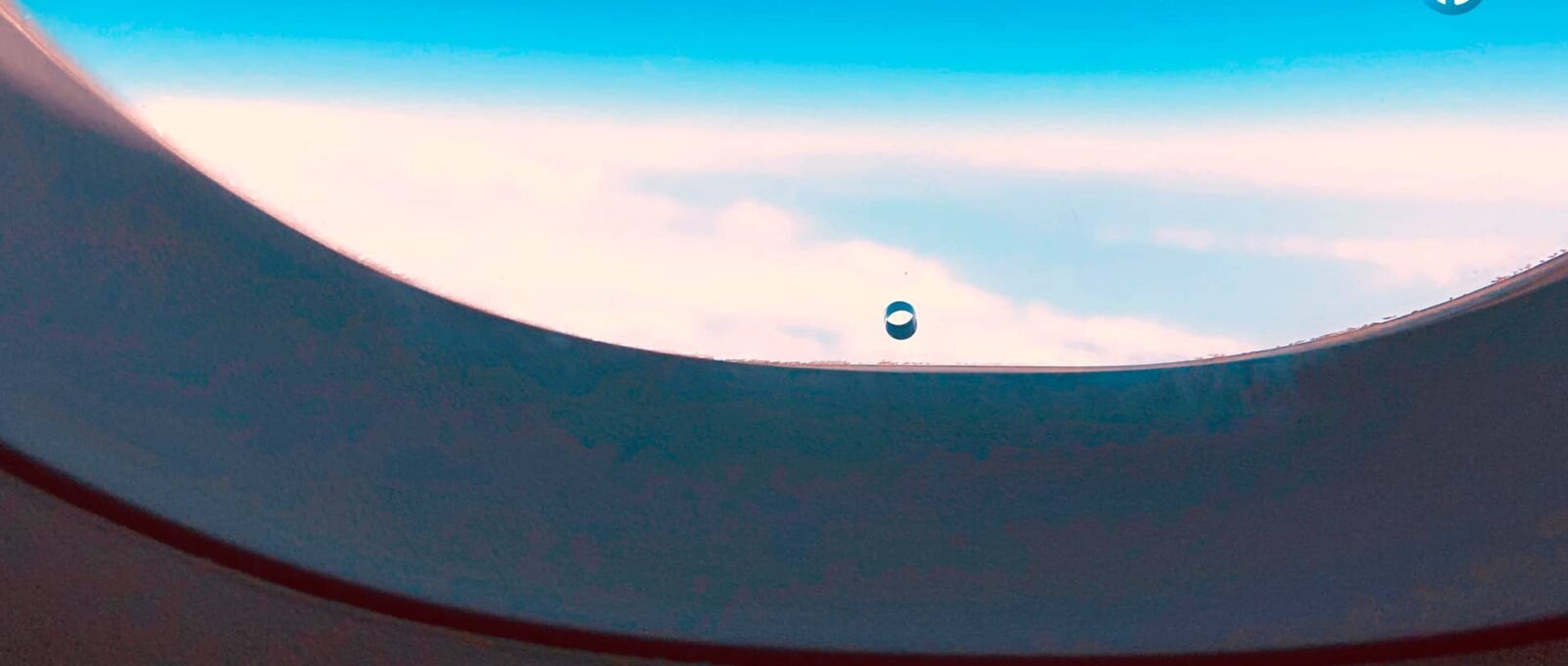 Vista a través de una ventana de avión mostrando un pequeño agujero circular cerca del borde de la ventana y un cielo azul con algunas nubes